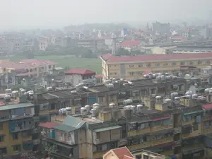 Đây là một bức ảnh chụp toàn cảnh một khu dân cư đông đúc tại một thành phố, có khả năng là Hà Nội, nhìn từ trên cao trong một ngày trời nhiều sương mù. Ở phía trước và trung tâm bức ảnh là các dãy nhà tập thể cũ, san sát nhau, tường sơn màu vàng đã ngả màu theo thời gian. Điểm đặc trưng nhất là trên nóc của hầu hết các tòa nhà này đều có rất nhiều bồn chứa nước bằng inox để dự trữ nước sinh hoạt. Trên một số bồn nước có thể đọc được chữ 'SƠN HÀ', là một thương hiệu nổi tiếng. Nhiều ban công của các căn hộ được cơi nới hoặc lắp thêm khung sắt bảo vệ (thường gọi là 'chuồng cọp'), và có thể thấy quần áo được phơi bên ngoài. Xen kẽ giữa các khu nhà tập thể là những ngôi nhà khác với mái ngói đỏ hoặc mái tôn xanh. Ở phía sau, có một tòa nhà lớn hơn, sơn màu vàng và có mái đỏ, trông giống một trường học hoặc một cơ quan hành chính. Giữa các khu nhà là một khoảng đất trống còn cây cỏ xanh. Phía xa, cảnh quan thành phố chìm dần trong màn sương, chỉ còn thấy hình bóng của các tòa nhà và các cột ăng-ten truyền hình.。（日本語訳）。これは、ある都市、おそらくハノイの密集した住宅地の全景を、霧の多い日に上空から撮影した写真です。写真の手前と中央には、古い集合住宅が密集して並んでおり、黄色に塗られた壁は時間の経過とともに色褪せています。いくつかの貯水タンクには、有名なブランドである「SƠN HÀ（ソンハ）」という文字が読み取れます。多くのアパートのバルコニーは増築されたり、鉄製の防犯柵（一般的に「虎の檻」と呼ばれます）が取り付けられたりしており、外に洗濯物が干されているのが見えます。集合住宅の間には、赤い瓦屋根や青いトタン屋根の他の家々が点在しています。後方には、黄色に塗られ赤い屋根をした、より大きな建物があり、学校か行政機関のように見えます。住宅街の間には、緑の草木が残っている空き地があります。遠くでは、都市の景観が霧の中に徐々に沈み、建物の影とテレビアンテナの柱だけが見えます。