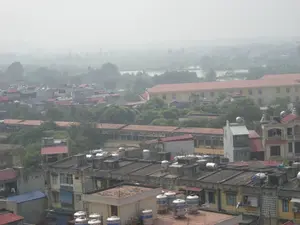 Bức ảnh chụp một quang cảnh từ trên cao của một khu dân cư đông đúc, có khả năng là ở vùng ngoại ô Hà Nội, trong một ngày có nhiều sương mù. Ở phía trước và trung tâm là vô số các ngôi nhà kiểu nhà ống san sát nhau, với các mái nhà bằng bê tông. Trên các mái nhà này có rất nhiều bồn chứa nước bằng inox để dự trữ nước sinh hoạt. Xen kẽ giữa các tòa nhà là cây xanh. Ở phía xa hơn một chút, có thể nhận ra các công trình kiến trúc lớn hơn, bao gồm một dãy nhà dài, thấp tầng với mái ngói đỏ và một tòa nhà lớn hơn, nhiều tầng, sơn màu vàng, có thể là một trường học hoặc một tòa nhà công cộng. Phía sau cùng, cảnh quan chuyển sang không gian mở hơn với một vùng nước (hồ hoặc sông) và các khu vực cây xanh, có thể là đất nông nghiệp, bị che khuất một phần bởi lớp sương mù dày đặc. Do góc chụp từ xa, không thể quan sát rõ các hoạt động cụ thể của con người. Không có văn bản hay biển hiệu nào có thể đọc được.。（日本語訳）。写真は、霧の多い日に、おそらくハノイ郊外にある、人口が密集した住宅地を上空から撮影した光景です。手前と中央には、コンクリート製の屋根を持つ、チューブハウス型の家が無数に密集しています。これらの屋根の上には、生活用水を貯蔵するためのステンレス製の貯水タンクが多数あります。建物の間には緑の木々が点在しています。少し奥には、赤い瓦屋根の長い低層の建物の一列と、学校または公共の建物である可能性のある、黄色に塗られたより大きな多層階の建物を含む、より大きな建築物が認識できます。一番奥では、景観は水域（湖または川）と、農地である可能性のある緑地エリアがある、より開けた空間に変わりますが、濃い霧の層によって部分的に覆われています。遠くからの撮影角度のため、人々の具体的な活動をはっきりと観察することはできません。読み取れる文字や看板はありません。