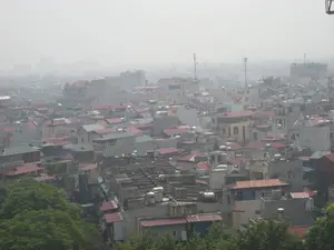 Toàn cảnh một khu vực đô thị đông đúc, có thể là Hà Nội, được chụp từ trên cao. Khung cảnh cho thấy vô số các tòa nhà cao tầng và nhà ống san sát nhau. Các mái nhà có nhiều màu sắc khác nhau, chủ yếu là mái ngói đỏ, mái tôn màu xanh lá cây và màu xám. Trên nóc của nhiều ngôi nhà có thể thấy rõ các bồn chứa nước bằng inox. Một vài cột ăng-ten thu phát sóng viễn thông nhô cao lên trên các tòa nhà. Bầu không khí mờ mịt trong sương mù hoặc khói bụi, che khuất các công trình ở phía xa. Phía dưới tiền cảnh là những tán cây xanh rậm rạp. Không có chữ viết hay biển hiệu nào có thể nhận dạng được. Do góc chụp từ xa, không thể quan sát được hoạt động cụ thể của con người.。（日本語訳）。上空から撮影された、ハノイと思われる人口密度の高い都市部の一帯の全景。屋根は様々な色をしており、主に赤瓦、緑や灰色のトタン屋根である。いくつかの通信送受信アンテナが建物のさらに上に突き出ている。大気は霧やスモッグでかすんでおり、遠くの建物を覆い隠している。前景の下部には、緑豊かな木々の葉が茂っている。識別できる文字や看板はない。遠くからの撮影角度のため、人々の具体的な活動を観察することはできない。