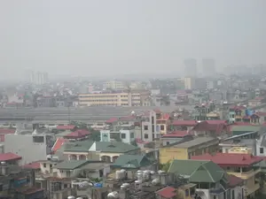 Bức ảnh chụp toàn cảnh một khu vực đô thị đông đúc, có thể là Hà Nội, nhìn từ trên cao. Bầu trời có màu xám trắng do sương mù hoặc ô nhiễm không khí dày đặc, làm hạn chế tầm nhìn ra xa. Ở phía trước là vô số các ngôi nhà dân thấp tầng san sát nhau, với các mái nhà có màu sắc đa dạng như đỏ và xanh lá. Trên nóc của nhiều ngôi nhà có lắp đặt các bồn chứa nước bằng inox, một hình ảnh quen thuộc tại các thành phố ở Việt Nam. Ở giữa bức ảnh, có một công trình kiến trúc dài với mái che màu xám, trông giống như một nhà ga xe lửa hoặc một khu chợ lớn có mái che. Ở phía xa trong hậu cảnh, lờ mờ hiện ra hình bóng của các tòa nhà cao tầng và chung cư. Do góc chụp từ xa và chất lượng hình ảnh, không thể thấy rõ hoạt động của con người hay bất kỳ văn bản nào trên các tòa nhà.。（日本語訳）。写真は、おそらくハノイと思われる、人口の密集した都市部を上空から撮影した全景です。空は濃い霧または大気汚染により白みがかった灰色で、遠くの視界を遮っています。手前には、赤や緑など様々な色の屋根を持つ低層の民家が無数に密集しています。多くの家の屋上にはステンレス製の貯水タンクが設置されており、これはベトナムの都市ではおなじみの光景です。写真の中央には、灰色の屋根を持つ長い建造物があり、駅または屋根付きの大きな市場のように見えます。遠くの背景には、高層ビルや集合住宅の影がぼんやりと現れています。遠くからの撮影角度と画質のため、人々の活動や建物上のいかなる文字もはっきりと見ることはできません。