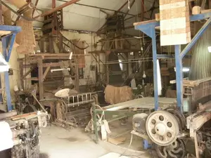 Đây là khung cảnh bên trong một xưởng dệt vải, có thể là một cơ sở sản xuất tại một làng nghề truyền thống. Không gian chứa đầy các máy dệt (khung cửi) lớn và phức tạp, được làm từ cả gỗ và kim loại. Các máy móc này trông khá cũ, bao gồm nhiều bộ phận cơ khí như bánh xe, bánh răng, động cơ điện và hệ thống dây chằng chịt. Mặc dù không có người nào xuất hiện, nhưng các máy dệt được bố trí như đang trong quá trình hoạt động sản xuất. Trên một máy dệt ở phía trước, các sợi chỉ màu trắng được căng sẵn. Ánh sáng trong xưởng được cung cấp bởi các bóng đèn tròn treo từ trần nhà có kết cấu dầm và mái tôn. Trên một chiếc máy ở phía sau, có một vật thể tròn, dẹt, trông giống một chiếc nón. Không có văn bản hay chữ viết nào có thể nhận dạng được trong ảnh.。（日本語訳）。これは織物工場の内部の光景で、伝統工芸村の生産施設かもしれません。空間は、木と金属の両方で作られた、大きくて複雑な織機（機織り機）でいっぱいです。これらの機械はかなり古く見え、車輪、歯車、電動モーター、そして複雑に絡み合ったワイヤーシステムなど、多くの機械部品で構成されています。人は誰も写っていませんが、織機は生産活動の最中であるかのように配置されています。手前にある一台の織機には、白い糸が張られています。工場内の照明は、梁とトタン屋根の構造を持つ天井から吊るされた丸い電球によって供給されています。写真の中には、認識できるテキストや文字はありません。