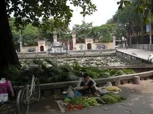 Bức ảnh chụp một khung cảnh tại Hà Nội. Ở tiền cảnh, một người phụ nữ đang ngồi bán rau trên vỉa hè dưới bóng một cây lớn. Cô bày bán nhiều loại rau củ như cà chua, dưa chuột và các loại rau xanh khác trên một tấm bạt. Bên cạnh cô có một chiếc cân đĩa nhỏ và một chiếc xe đạp cũ. Gần đó còn có một ma-nơ-canh trưng bày áo.。Hoạt động mua bán diễn ra bên cạnh một hồ nước có nhiều lá sen và hoa súng. Phía bên kia hồ là một công trình kiến trúc cổ, dường như là một ngôi đền. Công trình này có cổng, các cột trụ được trang trí và một bức tường bao quanh. Trên tường có các bức tranh vẽ những con voi. Phía trên cổng chính có khắc các ký tự chữ Hán nhưng không thể đọc rõ. Trong hậu cảnh, hai người đang đi bộ trên con đường ven hồ.。（日本語訳）。ハノイのある風景を写した写真。前景には、一人の女性が大きな木の陰の歩道で野菜を売って座っている。彼女はシートの上に、トマト、きゅうり、その他の青野菜といった多くの種類の野菜を並べて売っている。彼女の隣には、小さな皿秤と古い自転車がある。その近くには、服を展示しているマネキンもある。売買は、多くの蓮の葉と睡蓮がある池の隣で行われている。池の向こう側には、寺院と思われる古い建築物がある。この建物には門、装飾された柱、そして周囲を囲む壁がある。壁には象を描いた絵がある。正門の上には漢字が刻まれているが、はっきりと読むことはできない。背景では、二人の人が池沿いの道を歩いている。