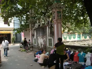 Trên mái hiên màu vàng của một cửa hàng ở bên trái có dòng chữ, một phần có thể đọc được là 'CẮT TÓC & GỘI ĐẦU', cho thấy đây có thể là một tiệm làm tóc. Các chữ khác bị mờ và không thể đọc rõ.。Bối cảnh là một con phố hoặc ngõ nhỏ dưới bóng cây xanh. Bên phải là một bức tường và cổng của một công trình kiến trúc cổ, có thể là đình hoặc chùa, với các cột trụ được trang trí hoa văn công phu. Bên cạnh bức tường dường như có một ao hoặc hồ nhỏ. Hoạt động chính trong ảnh là buôn bán trên vỉa hè. Một nhóm phụ nữ đang bày bán quần áo, với hàng hóa được đặt trên tấm bạt và trưng bày trên các ma-nơ-canh. Đây là một hình thức chợ cóc hoặc bán hàng rong. Ở giữa đường, một người khác đang bán trái cây (trông giống quả bưởi) trên một chiếc xe đạp. Có người đang đi bộ và người khác đang dừng xe máy gần người bán trái cây. Các vật thể có trong ảnh bao gồm xe máy, xe đạp, ma-nơ-canh, quần áo và trái cây.。（日本語訳）。左側にある店の黄色い軒先には文字があり、その一部は「CẮT TÓC & GỘI ĐẦU」（ヘアカット＆シャンプー）と読め、ここが美容院である可能性を示しています。他の文字はぼやけていて、はっきりと読むことはできません。背景は、緑の木陰にある通りまたは小さな路地です。右側には、古い建築物の壁と門があり、それはおそらく đình（村の集会所）か chùa（寺）で、柱には精巧な模様が装飾されています。壁の隣には、小さな池か湖があるようです。写真の中での主な活動は、歩道での商売です。女性たちのグループが服を売っており、商品はシートの上に置かれ、マネキンに展示されています。これは、chợ cóc（路上市場）または露天販売の一形態です。歩いている人もいれば、果物売りの近くでバイクを止めている人もいます。写真に写っている物には、バイク、自転車、マネキン、服、果物が含まれます。