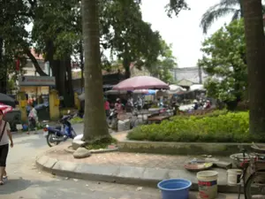 Trên một tòa nhà màu vàng có tấm biển ghi chữ 'BƯU ĐIỆN VĂN HÓA XÃ PHÚ...', phần chữ cuối không đọc được rõ. Khung cảnh là một khu chợ ngoài trời, có thể là một chợ cóc, họp ven một con đường nhựa. Nhiều người bán hàng ngồi dưới những chiếc ô lớn để che nắng, bày bán các mặt hàng trên sạp hoặc dưới đất. Mọi người đang đi lại mua sắm. Có thể thấy một người ở phía xa đang đội nón lá. Nhiều xe máy được dựng gần đó. Cảnh vật xung quanh có nhiều cây xanh, bao gồm cả cây cau, và một bồn cây nhỏ có viền bê tông. Ở phía trước bên phải, có một vài cái xô, chậu và một chiếc xe đạp cũ.。（日本語訳）。黄色い建物の上に「BƯU ĐIỆN VĂN HÓA XÃ PHÚ...」と書かれた看板があり、最後の文字ははっきりと読めない。風景は屋外市場で、アスファルトの道の脇で開かれている、おそらくは路上市場（chợ cóc）である。多くの売り手が日差しを避けるために大きな傘の下に座り、屋台の上や地面に商品を並べて売っている。人々が行き来しながら買い物をしている。多くのバイクがその近くに停められている。周りの景色には、ビンロウジュを含む多くの緑の木々や、コンクリートで縁取られた小さな植え込みがある。右前景には、いくつかのバケツや鉢、そして一台の古い自転車がある。