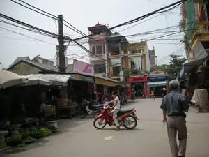 Bức ảnh ghi lại một khung cảnh trên một con phố nhỏ, có vẻ như là một khu chợ địa phương ở Việt Nam, có thể là Hà Nội.。**Chữ viết trên biển hiệu:**。*   Một cửa hàng có biển hiệu màu đỏ với chữ "HẢI PHÒNG".。*   Một cửa hàng khác có mái hiên sọc xanh và trắng, trên đó có biển hiệu ghi "PHÚC THÀNH" và dòng chữ nhỏ hơn "TỔ LỤA PHÚC THÀNH".。*   Ở phía trên một mái che, có một tấm biển quảng cáo lớn màu hồng với hình ảnh một người phụ nữ, một phần văn bản có thể đọc được là "...TUYÊN DỤNG CÙNG TỎA SÁNG...".。**Miêu tả cảnh vật và hoạt động:**。*   **Địa điểm:** Đây là một con phố với các cửa hàng và sạp hàng ở hai bên, mang dáng dấp của một khu chợ. Các tòa nhà là kiểu nhà ống nhiều tầng đặc trưng. Một đặc điểm nổi bật là mạng lưới dây điện và dây cáp chằng chịt vắt ngang qua đường và bám vào một cột điện lớn ở giữa.。*   **Hoạt động:** Một người phụ nữ đang lái một chiếc xe máy màu đỏ. Một người đàn ông lớn tuổi đang đi bộ trên đường. Bên lề trái, các sạp hàng có mái che đang bày bán các loại rau củ. Mọi người đang đi lại và có thể đang mua sắm tại các cửa hàng.。*   **Vật thể:** Hình ảnh có nhiều xe máy, các quầy hàng rong có mái che bằng bạt và ô, các rổ đựng rau củ, các tòa nhà ống, và một hệ thống cột điện và dây điện phức tạp. Ở phía xa, có thể nhận ra một người đang đội nón lá.。（日本語訳）。写真は、ベトナムの小さな通り、おそらくハノイの地元の市場と思われる風景を捉えています。**看板の文字：**。*   「HẢI PHÒNG」という文字が書かれた赤い看板のある店。*   青と白の縞模様の軒先テントがある別の店。その上には「PHÚC THÀNH」と書かれた看板と、より小さな文字で「TỔ LỤA PHÚC THÀNH」と書かれています。*   ある日よけの上には、女性の写真が載った大きなピンク色の広告看板があり、読み取れるテキストの一部は「...TUYÊN DỤNG CÙNG TỎA SÁNG...」です。**風景と活動の描写：**。*   **場所：** これは両側に店や屋台が並ぶ通りで、市場の様相を呈しています。建物は特徴的な多層階のチューブハウス（間口が狭く奥行きが深い家）です。際立った特徴は、通りを横切り、中央の大きな電柱に絡みつく、びっしりと張り巡らされた電線とケーブルの網です。*   **活動：** 一人の女性が赤いバイクを運転しています。年配の男性が道を歩いています。左側では、日よけのある屋台で様々な野菜が売られています。人々が行き交い、店で買い物をしている可能性があります。遠くには、ノンラー（ベトナムの伝統的な円錐形の帽子）をかぶっている人が一人見えます。