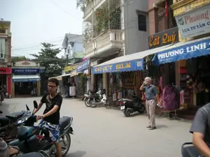 Bối cảnh là một con phố buôn bán ở Việt Nam, có thể là làng lụa Vạn Phúc ở Hà Đông, Hà Nội, dựa trên các biển hiệu. Các dòng chữ trên biển hiệu của các cửa hàng bao gồm: "PHÚC THÀNH", "TỔ LỤA PHÚC THÀNH", "Cát Quý", "CÁT QUÝ SILK", "PHƯƠNG LINH", và một biển hiệu khác ghi "LỤA TƠ TẰM" cùng địa chỉ "ĐC: VẠN PHÚC - HÀ ĐÔNG". Con phố có nhiều ngôi nhà ống san sát nhau. Các cửa hàng ở tầng trệt có mái hiên màu xanh và bày bán các sản phẩm dệt may như quần áo và khăn quàng. Nhiều xe máy (xe gắn máy) đang đậu ven đường. Ở phía trước, một thanh niên đang ngồi trên xe máy. Ở phía sau, một người đàn ông lớn tuổi đang đứng bên đường và nói chuyện điện thoại. Khung cảnh thể hiện hoạt động buôn bán và sinh hoạt thường ngày trên một con phố làng nghề.。（日本語訳）。背景はベトナムのある商店街で、看板からハノイ市ハドン区のヴァンフック・シルク村である可能性があります。店の看板の文字には、「PHÚC THÀNH」、「TỔ LỤA PHÚC THÀNH」、「Cát Quý」、「CÁT QUÝ SILK」、「PHƯƠNG LINH」が含まれており、また別の看板には「LỤA TƠ TẰM」という文字と「住所：ヴァンフック - ハドン」という住所が書かれています。通りには多くのチューブハウスが密集しています。1階の店には緑色の軒先テントがあり、衣類やスカーフなどの繊維製品が陳列・販売されています。多くのバイク（原動機付自転車）が道端に駐車されています。手前には、一人の若者がバイクに座っています。奥では、年配の男性が道端に立って電話で話しています。この光景は、伝統工芸村の通りにおける商売と日常生活の様子を表しています。