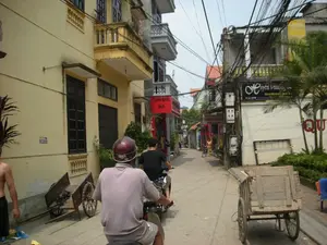 Bức ảnh chụp một con ngõ nhỏ ở Hà Nội. Hai bên đường là những ngôi nhà ống cao tầng, sơn màu vàng, với ban công. Khung cảnh đặc trưng với hệ thống dây điện và dây cáp chằng chịt phía trên. Hoạt động chính là giao thông hàng ngày, với những người đang đi xe máy. Một người đàn ông đội mũ bảo hiểm màu đỏ đang ngồi sau xe máy ở gần máy ảnh nhất. Ở lề đường, có một chiếc xe kéo tay bằng gỗ và một chiếc xe đẩy hai bánh khác. Có một số biển hiệu cửa hàng có thể nhìn thấy: một biển hiệu màu đỏ ghi "TUẤN KHẢI SILK", và một biển hiệu màu đen ghi "Huyền Phương". Trên bức tường vàng bên trái, có một số điện thoại được in: "091505000...". Xa hơn trong ngõ, một người phụ nữ đang đi bộ cùng với chiếc xe đạp.。（日本語訳）。ハノイの小さな路地を写した写真です。道の両側には、黄色に塗られたバルコニー付きの高層の ống（チューブ）型住宅が並んでいます。上部に張り巡らされた電線とケーブルのシステムが特徴的な光景です。主な活動は日常の交通で、バイクに乗っている人々がいます。カメラに最も近いバイクの後ろに、赤いヘルメットをかぶった男性が座っています。道端には、木製の手押し車と、もう一台の二輪の荷車があります。いくつかの店の看板が見えます。「TUẤN KHẢI SILK」と書かれた赤い看板と、「Huyền Phương」と書かれた黒い看板です。左側の黄色い壁には、「091505000...」という電話番号が印刷されています。路地のさらに奥では、女性が自転車と一緒に歩いています。