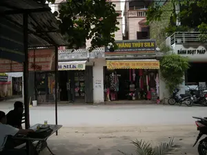 Đây là một khung cảnh đường phố tại một khu vực thương mại ở Việt Nam, có khả năng là một làng nghề chuyên về lụa ở gần Hà Nội. Con đường được trải nhựa và khá vắng vẻ.。Các thông tin văn bản có thể nhận dạng được trên các biển hiệu:。- Biển hiệu chính màu đen: 'THẮNG THỦY SILK', với dòng chữ nhỏ hơn bên dưới 'CHUYÊN BUÔN CÁC SẢN PHẨM LỤA TƠ TẰM CHẤT LƯỢNG CAO'.。- Mái hiên màu vàng của cùng cửa hàng: 'THẮNG THỦY SILK - LỤA TƠ TẰM TRUYỀN THỐNG'.。- Cửa hàng bên phải có biển hiệu 'BÁNH NGỌT PHÁP'.。- Cửa hàng bên trái có biển hiệu 'H SILK' và một biển hiệu màu đỏ bị che khuất một phần, chữ bị ngược, dường như là 'CỬA HÀNG BÁN BUÔN'.。Hoạt động và các vật thể trong ảnh:。- Địa điểm: Một con phố với các cửa hàng mặt tiền. Các tòa nhà có kiến trúc kiểu nhà phố, một số có ban công.。- Hoạt động: Các cửa hàng đang mở cửa kinh doanh. Cửa hàng lụa 'Thắng Thủy Silk' đang trưng bày nhiều sản phẩm như quần áo trên ma-nơ-canh, khăn quàng, và túi xách. Ở phía tiền cảnh, dưới một mái che lợp tôn, hai người đàn ông đang ngồi tại một chiếc bàn gỗ, có vẻ như đang trò chuyện và uống nước.。- Vật thể: Nhiều cửa hàng, biển hiệu, xe máy (hai chiếc đỗ trên vỉa hè và một phần của chiếc thứ ba ở bên phải), bàn ghế, ma-nơ-canh, các sản phẩm lụa, cây xanh.。（日本語訳）。これはベトナムのある商業地区の街の風景で、ハノイ近郊の絹織物専門の工芸村である可能性があります。道路は舗装されており、かなり閑散としています。看板から読み取れるテキスト情報：。- 黒いメイン看板：「THẮNG THỦY SILK」、その下にはより小さな文字で「高品質シルク製品専門卸売」。- 同じ店の黄色い庇：「THẮNG THỦY SILK - 伝統的なシルク」。- 右側の店には「BÁNH NGỌT PHÁP」（フランス菓子）という看板があります。- 左側の店には「H SILK」という看板と、一部が隠れ、文字が反転している赤い看板があり、それは「CỬA HÀNG BÁN BUÔN」（卸売店）のようです。写真の中の活動と物体：。- 場所：店舗が軒を連ねる通り。建物はタウンハウス様式の建築で、いくつかにはバルコニーがあります。- 活動：店は営業中です。シルク店「Thắng Thủy Silk」は、マネキンに着せられた服、スカーフ、ハンドバッグなど多くの商品を展示しています。前景では、トタン屋根の下で、二人の男性が木製のテーブルに座っており、おしゃべりをしたり飲み物を飲んだりしているようです。
