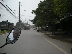 Bức ảnh chụp lại khung cảnh một con đường ở Việt Nam, có thể là ở khu vực ngoại ô Hà Nội. Góc chụp từ trên một chiếc xe máy đang di chuyển, với gương chiếu hậu ở phía trước bên trái phản chiếu hình ảnh một người đàn ông đeo kính. Con đường được trải nhựa và có nhiều phương tiện qua lại, bao gồm các xe tải ben lớn, ô tô con và xe máy. Bên trái đường là dải phân cách có trồng cây, nơi một chiếc xe tải đang đỗ và có một người ngồi trên thùng xe. Bên phải là vỉa hè rợp bóng cây xanh, nơi có các hàng quán nhỏ ven đường với một vài người đang ngồi, có thể là bán hàng. Nhiều xe máy khác đang đỗ trên vỉa hè. Bầu trời u ám và có rất nhiều dây điện, dây cáp chằng chịt trên các cột điện. Có chữ trên thân xe tải nhưng khó đọc, có thể nhận ra chữ 'CAT' trên một chiếc xe. Các biển hiệu của cửa hàng ven đường cũng không thể đọc được.。（日本語訳）。この写真はベトナムのある道路の風景を撮影したもので、おそらくハノイ郊外の地域でしょう。撮影アングルは走行中のバイクの上からで、左前方のバックミラーには眼鏡をかけた男性の姿が映っています。道路は舗装されており、大型ダンプカー、乗用車、バイクなど多くの車両が行き交っています。道路の左側には植樹された中央分離帯があり、そこには一台のトラックが停車し、荷台に人が一人座っています。右側は緑の木々が影を落とす歩道で、そこには道端の小さな店があり、数人が座っています。おそらく店の人でしょう。他にも多くのバイクが歩道に停められています。空は曇っており、電柱にはたくさんの電線やケーブルが複雑に張り巡らされています。トラックの車体には文字がありますが、読みづらく、一台の車に「CAT」という文字が認識できます。道端の店の看板も読み取ることができません。
