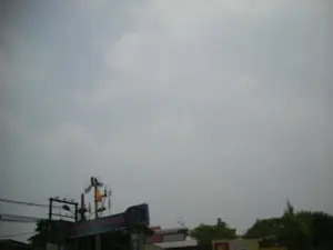 Bức ảnh chụp một khung cảnh đường phố dưới bầu trời u ám, nhiều mây. Ở phía dưới, có một tấm biển hiệu lớn màu xanh lam, chữ trên biển bị mờ nhưng có thể đọc được là 'LÀNG LỤA VẠN PHÚC'. Đây có thể là cổng chào hoặc biển chỉ dẫn vào Làng lụa Vạn Phúc ở Hà Nội. Bên cạnh tấm biển là một cột điện với hệ thống dây điện chằng chịt, đèn đường, và một lá cờ màu cam đang bay. Phía sau có thể thấy ngọn cây xanh và một phần mái nhà. Không có hoạt động cụ thể nào của con người được ghi lại trong ảnh.。（日本語訳）。写真は、どんよりとした曇り空の下の街の風景を写しています。下部には、大きな青い看板があり、看板の文字はぼやけていますが、「LÀNG LỤA VẠN PHÚC」と読み取れます。これは、ハノイにあるヴァンフック・シルク村へのウェルカムゲートまたは案内標識である可能性があります。看板の隣には、複雑に絡み合った電線、街灯、そしてはためくオレンジ色の旗が付いた電柱があります。後方には、緑の木々の梢と屋根の一部が見えます。写真には、特定の人の活動は記録されていません。