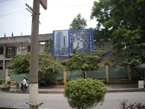 Trên một tấm biển quảng cáo lớn màu xanh lam có dòng chữ về một dự án xây dựng: 'KHU TỔ HỢP... TÒA THÁP THIÊN NIÊN KỶ HÀ TÂY'. Các dòng chữ nhỏ hơn bên dưới không thể đọc rõ. Bối cảnh là một con đường nhựa ở thành thị. Hai người (có vẻ là phụ nữ) đang đi chung một chiếc xe đạp trên đường. Trên vỉa hè, một người đàn ông đang đứng, và hai người khác đang ngồi nghỉ dưới bóng cây. Phía sau tấm biển quảng cáo là một tòa nhà hai tầng, trên nóc có cắm nhiều lá cờ với các màu đỏ, vàng, xanh và trắng. Nhiều cây xanh được trồng dọc theo vỉa hè. Một cột điện bê tông lớn nằm ở phía trước bên trái của khung hình.。（日本語訳）。青い大きな広告看板に、ある建設プロジェクトに関する文字が書かれている：「複合施設...ハタイ・ミレニアム・タワー」。その下のより小さな文字ははっきりと読み取れない。背景は都市部のアスファルトの道路である。二人（女性のようだ）が道路で一台の自転車に相乗りしている。歩道では、一人の男性が立っており、他の二人が木陰で座って休んでいる。歩道に沿って多くの緑の木が植えられている。フレームの左手前には、大きなコンクリートの電柱がある。