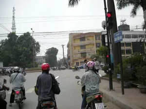 Bức ảnh chụp một cảnh giao thông tại một ngã tư ở Hà Nội. Có một biển báo đường phố màu xanh lam ghi chữ 'PHỐ CHU VĂN AN'. Nhiều người đang đi xe máy trên đường và một số đang dừng lại chờ đèn tín hiệu giao thông đang bật màu đỏ. Hầu hết mọi người đều đội mũ bảo hiểm; ở phía trước có thể thấy rõ một người đội mũ bảo hiểm màu đỏ, một người đội mũ màu hồng và một người đội mũ màu trắng. Xung quanh là các tòa nhà nhiều tầng, cây xanh và nhiều dây điện chằng chịt. Ở phía xa bên trái có một tháp ăng-ten viễn thông cao.。（日本語訳）。ハノイのある交差点の交通風景を写した写真です。「PHỐ CHU VĂN AN」と書かれた青い道路標識があります。多くの人が道路でバイクに乗っており、何人かは赤信号で止まって待っています。ほとんどの人がヘルメットをかぶっています。前方には、赤いヘルメットをかぶった人、ピンクのヘルメットをかぶった人、そして白いヘルメットをかぶった人がはっきりと見えます。周りには複数階建ての建物、緑の木々、そして多くの絡み合った電線があります。遠くの左側には、高い通信アンテナ塔があります。