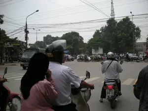 Cảnh chụp tại một ngã tư đường phố đông đúc, có thể là ở Hà Nội, vào một ngày trời u ám. Dòng phương tiện giao thông gồm rất nhiều xe máy, một vài chiếc ô tô và một chiếc xe tải đang dừng lại, có thể là để chờ đèn tín hiệu. Trên đường có vạch kẻ dành cho người đi bộ. Ở phía trước, một người đàn ông mặc áo sơ mi trắng đang lái xe máy, chở một người phụ nữ mặc áo hồng và trùm khăn đen ở phía sau. Hầu hết mọi người đi xe máy đều đội mũ bảo hiểm. Hai bên đường có cây cối xanh tươi. Phía trên là một mạng lưới dây điện và dây cáp chằng chịt. Ở phía xa có thể nhìn thấy một tháp viễn thông. Bên trái có một tấm biểu ngữ treo trên cây, trên đó có chữ nhưng chỉ đọc được một phần có thể là 'GẠO SẠCH KIẾN SƠN'.。（日本語訳）。曇りの日、おそらくハノイの、混雑した街の交差点で撮影された光景。交通の流れは非常に多くのバイク、数台の自動車、一台のトラックからなり、おそらく信号待ちで停止している。道路には歩行者用の横断歩道が引かれている。前方では、白いシャツを着た男性がバイクを運転しており、後ろにはピンクの服を着て黒いスカーフをかぶった女性を乗せている。バイクに乗っているほとんどの人がヘルメットをかぶっている。道路の両側には青々とした木々がある。上方には、電線とケーブルが複雑に絡み合った網のようになっている。左側には木に吊るされた横断幕があり、そこには文字が書かれているが、「GẠO SẠCH KIẾN SƠN」と思われる部分しか読み取れない。