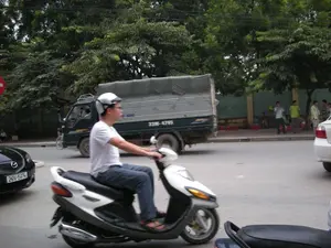 Cảnh đường phố tại một khu vực đô thị, có thể là Hà Nội. Một người đàn ông đội mũ bảo hiểm trắng, mặc áo phông trắng và quần jean đang điều khiển một chiếc xe máy tay ga trên đường nhựa. Cùng chiều với anh là một chiếc xe tải màu xanh đậm có mui bạt. Ở phía trước, có một phần của chiếc ô tô màu đen. Bên lề đường, trên vỉa hè, một nhóm người đàn ông đang tụ tập, một số đứng, một số ngồi trên những chiếc ghế nhựa nhỏ màu đỏ. Dọc theo con đường có nhiều cây xanh lớn. Một biển báo giao thông hình tròn màu đỏ và trắng bị che khuất một phần ở phía bên trái của hình ảnh.。（日本語訳）。ある都市部の街の風景、おそらくハノイだろう。白いヘルメットをかぶり、白いTシャツとジーンズを着用した男性が、アスファルトの道路でスクーターを運転している。彼と同じ方向には、幌付きの濃い青色のトラックが一台いる。道端の歩道では、男性のグループが集まっており、立っている人もいれば、小さな赤いプラスチックの椅子に座っている人もいる。道に沿って大きな緑の木がたくさんある。画像の左側で、赤と白の丸い交通標識が一部隠れている。