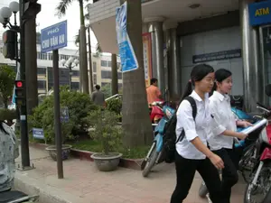 Chữ viết được ghi lại:。- Biển báo đường phố màu xanh lam ghi: "PHỐ CHU VĂN AN".。- Biển báo trên tòa nhà: "ATM".。- Dòng chữ trên cửa kính của tòa nhà: "KHÔNG SỬ DỤNG DỊCH VỤ BƯU ĐIỆN".。- Một biển báo nhỏ hơn ở khu vực cây xanh ghi: "CẤM BÁN HÀNG VÀ ĐỖ XE".。Mô tả cảnh vật và hoạt động:。Bức ảnh chụp một cảnh đường phố ở Hà Nội, trên vỉa hè của Phố Chu Văn An. Hai nữ sinh mặc đồng phục áo sơ mi trắng và quần tối màu đang đi bộ trên vỉa hè lát gạch. Một nữ sinh đeo kính và mang ba lô, trong khi người kia cầm một tập tài liệu. Họ có vẻ đang trên đường đến trường hoặc từ trường về nhà. Xung quanh có nhiều xe máy đang đỗ. Ở phía sau là một tòa nhà hiện đại với các cột trụ lớn, có một máy ATM. Một cột đèn giao thông đang hiển thị đèn đỏ. Cây xanh và các chậu cây cảnh được đặt dọc theo vỉa hè, tạo nên một khung cảnh đô thị điển hình.。（日本語訳）。記録された文字：。- 青い道路標識の表記：「PHỐ CHU VĂN AN」（チュー・ヴァン・アン通り）。- 建物にある看板：「ATM」。- 建物のガラスドアの文字：「KHÔNG SỬ DỤNG DỊCH VỤ BƯU ĐIỆN」（郵便サービスは利用しないでください）。- 緑地エリアにある小さな看板の表記：「CẤM BÁN HÀNG VÀ ĐỖ XE」（物販および駐車禁止）。風景と活動の説明：。写真はハノイの街角、チュー・ヴァン・アン通りの歩道で撮影されたものである。白いシャツと暗い色のズボンの制服を着た2人の女子生徒が、タイル張りの歩道を歩いている。一人の女子生徒は眼鏡をかけ、バックパックを背負っており、もう一人は書類の束を持っている。彼女たちは登校中か下校中のようだ。周りには多くのバイクが駐車している。信号機の柱は赤信号を表示している。緑の木々や鉢植えの観葉植物が歩道に沿って置かれ、典型的な都市の風景を作り出している。