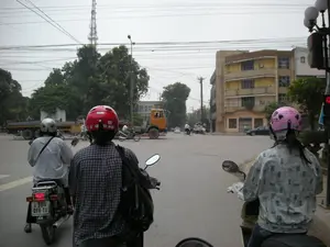 Bức ảnh chụp lại một cảnh giao thông tại một ngã tư ở Hà Nội. Góc nhìn từ phía sau một người đang ngồi trên xe máy. Nhiều người đi xe máy đang dừng chờ đèn đỏ. Tất cả những người đi xe máy đều đội mũ bảo hiểm: người ở giữa đội mũ màu đỏ, người bên trái đội mũ màu trắng, và một người phụ nữ bên phải đội mũ màu hồng và mặc áo chống nắng. Ở phía xa có một chiếc xe tải màu vàng, các phương tiện khác, những tòa nhà nhiều tầng, cây cối và một tháp ăng-ten viễn thông. Hệ thống dây điện chằng chịt vắt ngang trên đường phố.。（日本語訳）。写真はハノイのある交差点の交通風景を撮影したものです。バイクに乗っている人の後ろからの視点です。多くのバイクに乗った人々が赤信号で停止しています。バイクに乗っている人は全員ヘルメットを着用しています。中央の人は赤いヘルメット、左側の人は白いヘルメット、そして右側の女性はピンクのヘルメットをかぶり、日焼け防止の上着を着ています。遠くには黄色いトラック、その他の車両、高層ビル、木々、そして通信アンテナ塔が見えます。複雑に絡み合った電線が通りを横切っています。