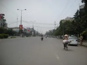 Bối cảnh là một con đường lớn, có nhiều làn xe ở thành phố, có thể là Hà Nội. Con đường có dải phân cách trồng cây bụi ở giữa và được trang bị hệ thống cột đèn đường. Hai bên đường là các tòa nhà nhiều tầng và cây xanh. Bầu trời u ám.。Hoạt động chính là cảnh sinh hoạt và giao thông trên đường phố. Nhiều người đang di chuyển bằng xe máy và xe đạp. Ở phía trước bên phải, một người đội nón lá đang đạp một chiếc xe đạp chở rất nhiều hàng hóa cồng kềnh được bọc trong tấm bạt hoặc bao tải. Gần đó, một chiếc ô tô taxi màu trắng đang đỗ bên lề đường, và một người phụ nữ khác cũng đội nón lá đang đứng bên cạnh. Ở phía xa hơn, có thể thấy nhiều xe máy khác đang lưu thông.。Về chữ viết, trên kính sau của chiếc xe taxi có chữ 'TAXI' và một số điện thoại nhưng không đọc được rõ. Dọc theo dải phân cách, trên các cột đèn có treo các tấm cờ phướn dọc màu đỏ, tuy nhiên chữ viết trên đó quá mờ để có thể đọc được. Ở phía xa cũng có các biển quảng cáo nhưng không thể nhận dạng nội dung.。（日本語訳）。背景は、都市の多くの車線がある大通りで、おそらくハノイでしょう。通りには中央に低木が植えられた中央分離帯があり、街灯システムが設置されています。通りの両側には高層ビルと緑の木々があります。空は曇っています。主な活動は、通りでの生活と交通の光景です。多くの人々がバイクや自転車で移動しています。右手前では、ノンラーをかぶった人が、防水シートか麻袋に包まれたたくさんのかさばる荷物を積んだ自転車をこいでいます。その近くでは、一台の白いタクシーが路肩に停車しており、同じくノンラーをかぶった別の女性がそのそばに立っています。さらに遠くには、他の多くのバイクが通行しているのが見えます。中央分離帯に沿って、街灯の柱には赤い縦長の幟旗が掲げられていますが、その上の文字はぼやけすぎていて読むことができません。遠くにも広告看板がありますが、内容を識別することはできません。