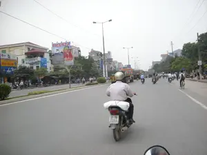 Đây là hình ảnh chụp một con đường lớn, có nhiều làn xe ở Hà Nội. Khung cảnh giao thông tấp nập với nhiều người đang di chuyển, chủ yếu bằng xe máy. Ở phía trước, một người đàn ông đội mũ bảo hiểm đang lái xe máy, phía sau yên xe có chở một bọc hàng màu trắng. Trên đường còn có các phương tiện khác như xe tải và xe đạp. Hai bên đường là những dãy nhà cao tầng san sát, có nhiều cửa hàng kinh doanh. Có rất nhiều biển hiệu và biển quảng cáo có thể nhìn thấy, bao gồm: một biển quảng cáo lớn trên nóc nhà cho thương hiệu "PICENZA" với dòng chữ "BÌNH NƯỚC NÓNG", các biển hiệu cửa hàng "LG", "ĐIỆN TỬ - ĐIỆN LẠNH", "Âm Thanh", và "SAMSUNG". Ngoài ra còn có một pano cổ động lớn màu đỏ có hình Chủ tịch Hồ Chí Minh và các dòng chữ như "ĐẢNG CỘNG SẢN VIỆT NAM QUANG VINH MUÔN NĂM。" và "CHỦ TỊCH HỒ CHÍ MINH VĨ ĐẠI SỐNG MÃI TRONG SỰ NGHIỆP CỦA CHÚNG TA". Trên đường có các cột đèn chiếu sáng và hệ thống dây điện chằng chịt. Bầu trời có vẻ u ám.。（日本語訳）。これはハノイの多車線ある大きな道路を撮影した写真です。交通は混雑しており、多くの人々が主にバイクで移動しています。前方では、ヘルメットをかぶった男性がバイクを運転しており、後部座席には白い荷物を載せています。道路にはトラックや自転車などの他の車両もあります。道路の両側には高層ビルが立ち並び、多くの商店が営業しています。多くの看板や広告が見え、それには「BÌNH NƯỚC NÓNG」（温水器）という文字が入った「PICENZA」ブランドの屋上の大きな広告看板、「LG」、「ĐIỆN TỬ - ĐIỆN LẠNH」（電子-冷凍電気）、「Âm Thanh」（音響）、そして「SAMSUNG」の店舗看板などが含まれます。その他、ホー・チ・ミン主席の肖像と「ĐẢNG CỘNG SẢN VIỆT NAM QUANG VINH MUÔN NĂM。」（ベトナム共産党万歳。）や「CHỦ TỊCH HỒ CHÍ MINH VĨ ĐẠI SỐNG MÃI TRONG SỰ NGHIỆP CỦA CHÚNG TA」（偉大なるホー・チ・ミン主席は我々の事業の中に永遠に生き続ける）といったスローガンが書かれた大きな赤いプロパガンダパネルもあります。道路には街灯の柱と、複雑に絡み合った電線システムがあります。空は曇っているようです。