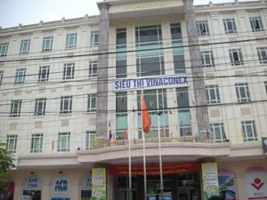 Bức ảnh chụp một tòa nhà thương mại lớn, nhiều tầng với kiến trúc hiện đại. Phía trên cùng của tòa nhà có dòng chữ 'VINACONEX'. Một biển hiệu lớn màu xanh dương ở mặt tiền ghi 'SIÊU THỊ VINACONEX'. Tòa nhà được sơn màu kem, có nhiều cửa sổ và các cột trụ lớn. Phía trước tòa nhà có rất nhiều dây điện chằng chịt. Một lá cờ Việt Nam màu đỏ sao vàng và nhiều cờ đuôi nheo đủ màu sắc được treo để trang trí.。Ở tầng trệt, có thể thấy logo của các ngân hàng như 'ACB' và 'BIDV'. Một băng rôn lớn màu đỏ được treo ngang ban công với nội dung: 'NHIỆT LIỆT CHÀO MỪNG LỄ KỶ NIỆM 20 NĂM NGÀY THÀNH LẬP TỔNG CÔNG TY VINACONEX (27/09/1988 - 27/09/2008)', cho thấy đang có một sự kiện kỷ niệm đang diễn ra. Bên phải có một băng rôn quảng cáo dọc ghi 'Cơ hội trúng lớn tại Hà Đông plaza' với các giải thưởng được liệt kê như 'MÁY GIẶT LG', 'TIVI SONY', 'BẾP GAS', 'NỒI CƠM ĐIỆN'. Khung cảnh cho thấy đây là một trung tâm mua sắm hoặc tòa nhà văn phòng phức hợp, đang được trang hoàng cho một dịp đặc biệt.。（日本語訳）。写真は、近代的な建築様式の大きな多層階の商業ビルを写しています。ビルの最上部には「VINACONEX」という文字があります。ファサードにある大きな青い看板には「SIÊU THỊ VINACONEX」（ビナコネックススーパーマーケット）と書かれています。ビルはクリーム色に塗られており、多くの窓と大きな柱があります。ビルの前には、たくさんの電線がごちゃごちゃと張り巡らされています。赤地に金星のベトナム国旗1枚と、色とりどりの多くの吹き流しが装飾のために掲げられています。1階には、「ACB」や「BIDV」といった銀行のロゴが見えます。バルコニーには「VINACONEX総公社設立20周年記念式典（1988年9月27日～2008年9月27日）を熱烈に歓迎します」という内容の大きな赤い横断幕が掲げられており、記念イベントが開催されていることを示しています。右側には「ハドンプラザで大当たりを掴むチャンス」と書かれた縦長の広告バナーがあり、「LG洗濯機」「ソニーのテレビ」「ガスコンロ」「炊飯器」などの賞品が記載されています。この光景は、ここがショッピングセンターまたは複合オフィスビルであり、特別な機会のために装飾されていることを示しています。