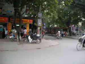 Bức ảnh chụp một góc phố ở Việt Nam, có thể là Hà Nội. Bên trái là một quán ăn có biển hiệu lớn màu đỏ và vàng, trên đó có ghi chữ "PHỞ ĐOÀN". Nhiều người đang tụ tập trước quán, có vẻ như đang mua đồ ăn hoặc nói chuyện. Trên vỉa hè có một vài chiếc xe máy đang đậu. Dưới lòng đường, một người phụ nữ đội nón lá đang đi xe đạp, và một người khác cũng đang đi xe đạp ở phía trước. Cảnh vật xung quanh có nhiều cây xanh và một cột điện. Khung cảnh tổng thể cho thấy một hoạt động sinh hoạt đời thường trên đường phố.。（日本語訳）。ベトナムのある街角を写した写真で、おそらくハノイでしょう。左側には赤と黄色の大きな看板を掲げた食堂があり、「PHỞ ĐOÀN」という文字が書かれています。店の前には多くの人が集まっており、食べ物を買ったり話したりしているようです。歩道には数台のバイクが停まっています。車道では、ノンラーをかぶった女性が自転車に乗っており、その前方にもう一人自転車に乗っている人がいます。周囲の景色には多くの緑の木々と一本の電柱があります。全体的な光景は、路上での日常的な生活の営みを示しています。