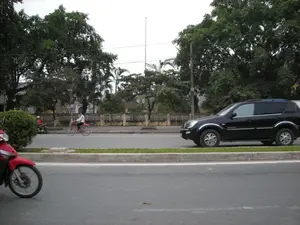 Đây là hình ảnh chụp một con đường giao thông ở khu vực đô thị. Con đường được trải nhựa và có một dải phân cách trồng cỏ ở giữa. Ở làn đường phía xa, một chiếc ô tô SUV màu đen đang di chuyển. Gần đó, một người mặc áo trắng đang đi xe đạp màu đỏ. Bên lề đường có rất nhiều cây xanh lớn và một hàng rào bê tông. Ở phía trước, góc dưới bên trái của ảnh là một phần của một chiếc xe máy màu đỏ. Không có chữ viết hay biển báo nào có thể đọc được trong ảnh.。（日本語訳）。これは都市部の交通路を撮影した写真です。道路は舗装されており、中央には草が植えられた分離帯があります。遠くの車線では、黒いSUV車が走行しています。その近くでは、白い服を着た人が赤い自転車に乗っています。道端には多くの大きな緑の木々とコンクリートの柵があります。写真には読み取れる文字や標識はありません。