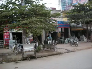 Quang cảnh một con phố ở Hà Nội với các cửa hàng kinh doanh vật liệu xây dựng. Trên vỉa hè, nhiều sản phẩm như khung cửa sổ, cửa ra vào bằng nhôm kính được trưng bày. Có một vài chiếc xe máy, một chiếc xe đạp gắn với xe kéo và một chiếc xe đẩy tay đang đỗ. Cây xanh lớn mọc dọc theo vỉa hè. Có một số biển hiệu của cửa hàng, trong đó một tấm biển màu xanh ở trung tâm có ghi chữ 'NHÔM KÍNH - SẮT - INOX'. Một tấm biển nhỏ khác dán trên thân cây có chữ 'CẤT GẠCH'. Một tấm biển màu đỏ bên trái có logo của Honda. Phía sau các cửa hàng là một tòa nhà cao tầng với một tấm biển quảng cáo lớn nhưng bị mờ.。（日本語訳）。ハノイのある通りの光景。建材を商う店々が並んでいる。歩道には、窓枠やアルミサッシ製のドアなど、多くの製品が展示されている。数台のバイク、リヤカーに繋がれた一台の自転車、そして一台の手押し車が停まっている。歩道に沿って大きな緑の木が生えている。いくつかの店の看板があり、そのうち中央にある青い看板には「NHÔM KÍNH - SẮT - INOX」という文字が書かれている。木の幹に貼られた別の小さな看板には「CẤT GẠCH」という文字がある。左側にある赤い看板にはホンダのロゴがある。店々の後ろには高層ビルがあり、大きな広告看板があるが、それはぼやけている。