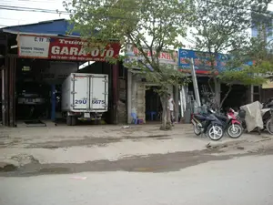 Bản ghi các thông tin trên biển hiệu:。- Biển hiệu chính màu đỏ và trắng của một gara ô tô: "C.TY TNHH Trường GARA Ô TÔ", chuyên "ĐẠI TU MÁY - GẦM - ĐIỆN LẠNH - SƠN GÒ".。- Một biển hiệu nhỏ hơn bên trái ghi "QUANG TRUNG" và số điện thoại "ĐT: 034. 514744".。- Một biển hiệu màu xanh ở cửa hàng bên cạnh bị cây che khuất phần lớn, không thể đọc rõ nội dung.。- Bên trong gara có một biển hiệu khác có chữ "PICO" nhưng phần còn lại không rõ.。Mô tả cảnh vật và hoạt động:。Đây là khung cảnh mặt tiền của một dãy cửa hàng ven đường, chủ yếu là các gara sửa chữa ô tô. Con đường phía trước không được trải nhựa hoàn toàn, có những mảng bê tông và đất lầy lội.。- Một gara lớn đang mở cửa hoạt động. Bên trong, một chiếc ô tô con đang được đặt trên cầu nâng, cho thấy hoạt động sửa chữa đang diễn ra. Một chiếc xe tải thùng màu trắng, hiệu ISUZU, đang đỗ ở lối vào của gara.。- Phía trước các cửa hàng, có nhiều xe máy đang đậu. Một người đàn ông mặc áo ba lỗ màu trắng đang đứng gần một trong hai cái cây xanh lớn ở phía trước. Cảnh vật cho thấy đây là một khu vực kinh doanh dịch vụ sửa chữa xe cộ.。（日本語訳）。看板情報の記録：。- ある自動車修理工場の赤と白のメイン看板：「C.TY TNHH Trường GARA Ô TÔ」、専門は「エンジンオーバーホール - シャーシ - エアコン - 板金塗装」。- 左側の小さな看板には「QUANG TRUNG」と電話番号「電話：034. 514744」と書かれている。- 隣の店の青い看板は、木によって大部分が隠されており、内容をはっきりと読み取ることはできない。- 修理工場の内部には「PICO」という文字のある別の看板があるが、残りの部分は不明瞭である。風景と活動の描写：。これは道端に並ぶ店舗の正面の光景であり、主に自動車修理工場である。正面の道路は完全には舗装されておらず、コンクリートとぬかるんだ土の部分がある。- 大きな修理工場が営業中である。内部では、乗用車1台がリフトに載せられており、修理作業が行われていることを示している。ISUZU製の白い箱型トラック1台が、修理工場の入り口に駐車している。- 店舗の前には、多くのバイクが停まっている。白いタンクトップを着た男性が、正面にある2本の大きな緑の木の1本の近くに立っている。この風景は、ここが車両修理サービス業の商業地域であることを示している。