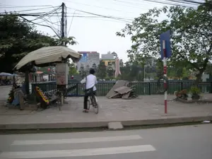 Bối cảnh là một con đường ở thành thị với vạch kẻ sang đường. Trên vỉa hè, một người mặc áo sơ mi trắng đang đi xe đạp. Bên trái, có một quầy hàng rong nhỏ được che bởi một chiếc ô lớn, dường như đang bán các vòng hoa. Gần đó là một cột điện với hệ thống dây điện chằng chịt. Ở giữa vỉa hè, có một đống vật liệu xây dựng và một cột biển báo giao thông sọc đỏ trắng, trên đó có treo một chiếc lốp xe cũ. Một con chó đang ở trên vỉa hè phía bên phải. Vỉa hè có lan can màu xanh lá cây, nhìn ra một khu vực công viên có cây cối và một tượng đài hình tên lửa ở phía xa. Phía sau là các tòa nhà. Có một số bảng hiệu và chữ viết trên một hộp điện gần quầy hàng nhưng không thể đọc rõ.。（日本語訳）。背景は、横断歩道のある都市の道路です。歩道の上では、白いシャツを着た人が自転車に乗っています。左側には、大きな傘で覆われた小さな屋台があり、花輪を売っているようです。その近くには、電線が複雑に絡み合った電柱があります。歩道の中央には、建設資材の山と、古いタイヤが吊るされた赤と白の縞模様の交通標識のポールがあります。右側の歩道には犬がいます。歩道には緑色の手すりがあり、木々や遠くにあるロケット型の記念碑がある公園エリアに面しています。後ろには建物があります。屋台の近くの配電箱にはいくつかの看板や文字がありますが、はっきりと読むことはできません。