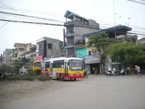 Bối cảnh là một con đường ở một khu vực đô thị tại Việt Nam, có thể là ở Hà Nội. Cảnh quan bao gồm các ngôi nhà ống nhiều tầng, đường sá và một bãi đất trống.。**Văn bản được ghi lại:**。*   Trên chiếc xe buýt màu vàng và đỏ: Phía trước có chữ "Tín Lợi". Bên hông xe có ghi tuyến đường "HÀ ĐÔNG - XUÂN MAI".。*   Biển hiệu cửa hàng: Một biển hiệu lớn màu xanh lam có chữ "KARAOKE".。*   Các biển hiệu khác: Có một biển quảng cáo màu đỏ phía sau xe buýt có chữ "SƠN..." nhưng phần còn lại bị che khuất. Một biển hiệu khác có logo "SYM" và chữ "XE MÁY". Một biển hiệu màu xanh trên tòa nhà phía xa có chữ "HOTEL" bị che một phần.。**Mô tả cảnh quan và hoạt động:**。*   **Địa điểm:** Đây là một con đường phố với những ngôi nhà ống nhiều tầng đặc trưng san sát nhau. Bên cạnh con đường nhựa là một bãi đất trống. Phía trên là một mạng lưới dây điện và cáp viễn thông chằng chịt.。*   **Hoạt động:** Một chiếc xe buýt đang đỗ bên lề đường, gần một bãi đất trống. Phía sau nó là một chiếc xe tải ben màu vàng. Một số người đang di chuyển bằng xe máy trên đường. Vài người khác đang ngồi trước cửa hàng karaoke. Ở phía bên phải, một người đang đi bộ.。*   **Vật thể:** Các phương tiện giao thông bao gồm xe buýt, xe tải, nhiều xe máy và xe đạp. Các tòa nhà là nhà ở kết hợp kinh doanh, nổi bật là cửa hàng karaoke. Cây xanh mọc ven đường và trước các ngôi nhà.。（日本語訳）。背景はベトナムのある都市部の道路で、おそらくハノイです。風景には、多層のチューブハウス、道路、そして空き地が含まれています。**記録されたテキスト：**。*   黄色と赤色のバス：前面に「Tín Lợi」の文字があります。車体の側面には「HÀ ĐÔNG - XUÂN MAI」の路線が書かれています。*   店の看板：大きな青い看板に「KARAOKE」の文字があります。*   その他の看板：バスの後ろに赤い広告看板があり、「SƠN...」という文字がありますが、残りの部分は隠されています。別の看板には「SYM」のロゴと「XE MÁY」の文字があります。遠くの建物にある青い看板には、一部が隠れた「HOTEL」の文字があります。**風景と活動の説明：**。*   **場所：**これは、特徴的な多層のチューブハウスが密集して建ち並ぶ街の通りです。アスファルト道路の隣には空き地があります。頭上には、電線と通信ケーブルの網が複雑に張り巡らされています。*   **活動：**一台のバスが空き地の近くの道端に停車しています。その後ろには黄色いダンプトラックがいます。何人かの人々がバイクで道路を移動しています。他の数人がカラオケ店の前に座っています。右側では、一人の人が歩いています。*   **物体：**交通手段には、バス、トラック、多くのバイク、自転車が含まれます。建物は住居兼店舗で、特にカラオケ店が目立ちます。緑の木が道端や家の前に生えています。