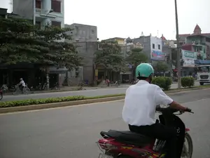 Bức ảnh chụp một cảnh đường phố ở Việt Nam, có thể là ở Hà Nội. Ở tiền cảnh, một người đàn ông mặc áo sơ mi trắng, đội mũ bảo hiểm màu xanh nhạt đang điều khiển một chiếc xe máy màu đỏ trên đường. Con đường được trải nhựa, có một dải phân cách cứng trồng cây bụi ở giữa.。Bên kia đường là một dãy nhà ống nhiều tầng, một số có ban công. Dưới tầng trệt là các cửa hàng kinh doanh. Có nhiều cây xanh lớn ven đường. Một người đội nón lá đang đi xe đạp trên vỉa hè hoặc sát lề đường. Nhiều xe máy khác đang đỗ trước các cửa hàng. Ở phía xa bên phải, có một chiếc xe tải màu trắng.。Trên các tòa nhà có một số biển hiệu quảng cáo. Một biển hiệu lớn có chữ 'caesar'. Một biển hiệu khác màu xanh dương có chữ 'Nghĩa - Nhung'. Một cửa hàng có biển hiệu dọc màu đen với chữ 'NOKIA'. Một biển hiệu khác có thể đọc được một phần là 'SỬA CHỮA XE MÁY'.。（日本語訳）。この写真はベトナムの街頭風景を撮影したもので、おそらくハノイでしょう。前景には、白いシャツを着て水色のヘルメットをかぶった男性が、道路で赤いバイクを運転しています。道路は舗装されており、中央には植え込みのある中央分離帯があります。道路の向こう側には、多層階の縦長の家が並んでおり、いくつかにはバルコニーが付いています。1階部分は店舗になっています。道路沿いには大きな緑の木がたくさんあります。ノンラー（菅笠）をかぶった人が、歩道または路肩で自転車に乗っています。他にも多くのバイクが店の前に駐車されています。右手の遠くには、白いトラックが1台あります。建物にはいくつかの広告看板があります。大きな看板には「caesar」という文字があります。別の青い看板には「Nghĩa - Nhung」という文字があります。ある店には「NOKIA」という文字が入った黒い縦長の看板があります。別の看板には、部分的に「SỬA CHỮA XE MÁY」（バイク修理）と読み取れます。