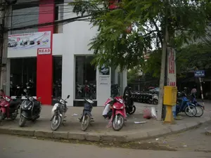 Chữ viết trên các biển hiệu và băng rôn:。- Băng rôn quảng cáo lớn treo trên tòa nhà: 'Sắm hàng hiệu, trúng về nhà ngay bây giờ.' cùng với logo 'CHELIMEX'.。- Biển hiệu màu đỏ của cửa hàng: 'HEAD' và 'Cửa hàng do Honda ủy nhiệm'.。- Biển báo đường phố màu xanh: 'NGÕ 12 ĐƯỜNG XUÂN THỦY'.。- Một biển quảng cáo dọc màu đỏ và trắng trên cột điện nhưng chữ bị mờ, không đọc rõ.。Mô tả cảnh vật và hoạt động:。Bức ảnh chụp một góc phố ở Hà Nội, tại ngã ba giao với Ngõ 12 đường Xuân Thủy. Địa điểm nổi bật là một cửa hàng bán xe máy của Honda (HEAD), có kiến trúc hiện đại với mặt tiền kính lớn và các mảng tường màu trắng, đỏ. Bên trong và trên vỉa hè phía trước cửa hàng trưng bày và đỗ rất nhiều xe máy (xe máy) mới để bán, bao gồm cả xe tay ga và xe số. Một người đang ngồi xổm bên cạnh một chiếc xe máy màu đỏ trên vỉa hè. Cảnh vật xung quanh bao gồm một cây xanh lớn, một cột điện, và một hòm thư công cộng màu vàng đặc trưng.。（日本語訳）。看板と横断幕の文字：。- 建物に掲げられた大きな広告横断幕：「ブランド品を買って、今すぐ家を当てよう。」、「CHELIMEX」のロゴと共に。- 店舗の赤い看板：「HEAD」と「ホンダ正規販売店」。- 青い道路標識：「NGÕ 12 ĐƯỜNG XUÂN THỦY」。- 電柱にある赤と白の縦型広告看板が一つあるが、文字がぼやけていてはっきりと読めない。風景と活動の説明：。写真はハノイのある街角、スアン・トゥイ通り12番路地との三叉路を撮影したものである。目立つ場所はホンダのバイク販売店（HEAD）で、大きなガラス張りのファサードと白と赤の壁面を持つ近代的な建築である。店内と店の前の歩道には、スクーターとマニュアル車の両方を含む、販売用の新しいバイク（バイク）が多数展示・駐車されている。一人の人が歩道で赤いバイクの隣にしゃがんでいる。周囲の風景には、大きな緑の木、一本の電柱、そして特徴的な黄色い公共郵便ポストが含まれている。