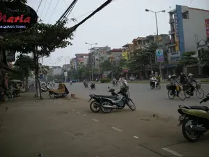 Đây là hình ảnh một con phố ở thành thị tại Việt Nam, có thể là Hà Nội, với nhiều hoạt động đời thường đang diễn ra.。**Chữ viết được ghi lại:**。*   Biển hiệu đèn LED màu đỏ ở góc trên bên trái có chữ "THẨM MỸ VIỆN Maria" và địa chỉ "34 QUANG TRUNG".。*   Trên một tòa nhà cao ở phía bên phải, có biển hiệu dọc màu xanh với chữ "SAMSUNG" và một biển hiệu khác ghi "NOKIA".。*   Trên vỉa hè có một tấm biển nhỏ màu đỏ ghi chữ "CẤM BÁN HÀNG".。*   Có nhiều biển hiệu quảng cáo khác trên các tòa nhà nhưng chữ viết quá nhỏ và mờ để có thể đọc được.。**Mô tả cảnh quan và hoạt động:**。*   **Địa điểm:** Một con phố đô thị sầm uất với các tòa nhà cao tầng san sát hai bên. Vỉa hè được lát gạch, nhưng có một số khu vực trông như đang được sửa chữa hoặc xuống cấp. Hệ thống dây điện và cáp viễn thông chằng chịt, vắt ngang qua đường là một đặc điểm rất nổi bật.。*   **Hoạt động:** Mọi người đang tham gia giao thông trên đường. Nhiều người đang đi xe máy (xe gắn máy). Có một nhóm người, trong đó có những phụ nữ đội nón lá, đang đi xe đạp. Một cửa hàng ở bên lề đường bên trái đang mở và có người đứng bán hàng.。*   **Vật thể:**。*   **Phương tiện:** Rất nhiều xe máy đang lưu thông trên đường và đỗ trên vỉa hè. Một chiếc xe máy được che phủ bằng một tấm bạt màu nâu. Ngoài ra còn có một vài chiếc xe đạp.。*   **Con người:** Người dân địa phương đang di chuyển, một số phụ nữ đội nón lá truyền thống.。*   **Công trình:** Các tòa nhà là sự kết hợp giữa nhà ở và mặt bằng kinh doanh, với nhiều cửa hàng và biển quảng cáo.。（日本語訳）。これはベトナムの都市部、おそらくハノイの街角の画像で、多くの日常的な活動が行われています。**記録された文字：**。*   左上の赤いLED看板には「THẨM MỸ VIỆN Maria」（マリア美容院）という文字と「34 QUANG TRUNG」という住所があります。*   右側の高い建物には、「SAMSUNG」と書かれた青い縦長の看板と、「NOKIA」と書かれた別の看板があります。*   歩道には「CẤM BÁN HÀNG」（商売禁止）と書かれた小さな赤い看板があります。*   建物には他にも多くの広告看板がありますが、文字が小さすぎてぼやけているため読むことができません。**景観と活動の説明：**。*   **場所：** 両側に高層ビルが立ち並ぶ、賑やかな都会の通り。歩道はタイルで舗装されていますが、一部は修理中または老朽化しているように見えます。複雑に絡み合い、道を横切る電線や通信ケーブルのシステムが非常に際立った特徴です。*   **活動：** 人々が道路で交通に参加しています。多くの人がバイク（原動機付自転車）に乗っています。ノンラーをかぶった女性を含む一団が自転車に乗っています。左側の路肩にある店が開いており、店員が立っています。*   **物体：**。*   **乗り物：** 非常に多くのバイクが道路を走行し、歩道に駐車しています。1台のバイクは茶色のシートで覆われています。その他に数台の自転車もあります。*   **人々：** 地元の人々が移動しており、一部の女性は伝統的なノンラーをかぶっています。*   **建造物：** 建物は住居と商業スペースが組み合わさったもので、多くの店舗や広告看板があります。