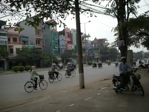 Bức ảnh chụp một cảnh đường phố ở Việt Nam, có thể là Hà Nội, với nhiều phương tiện giao thông đang di chuyển. Dọc bên đường là những dãy nhà ống nhiều tầng, sơn màu sắc sặc sỡ, đặc trưng của đô thị Việt Nam. Các ngôi nhà này dường như vừa là nhà ở, vừa là cửa hàng kinh doanh. Hoạt động chính là mọi người đang đi lại trên đường bằng xe đạp và xe máy. Ở phía trước, có ba người phụ nữ đang đạp xe, hai trong số họ đội nón lá truyền thống. Một chiếc xe đạp có giỏ hàng ở phía sau. Một người đàn ông đội mũ bảo hiểm đang ngồi trên xe máy ở lề đường. Cảnh vật bao gồm xe đạp, xe máy (xe máy), nón lá, nhà ống, cây cối ven đường và một cột điện lớn với hệ thống dây điện chằng chịt phía trên. Các thông tin văn bản có thể đọc được bao gồm biển hiệu "Ericsson", "NOKIA" trên các tòa nhà, một biển hiệu dọc có thể là "NHÀ NGHỈ" và một tấm biển viết tay treo trên cây ghi "ÁO DA BÁN".。（日本語訳）。写真は、多くの乗り物が移動しているベトナムの、おそらくハノイの街の風景を写したものです。道沿いには、ベトナムの都市に特徴的な、鮮やかな色で塗られた多層階のチューブハウスが並んでいます。これらの家は住居でもあり、商店でもあるようです。主な活動は、人々が自転車やバイクで路上を行き来していることです。手前には、自転車に乗っている3人の女性がおり、そのうちの2人は伝統的なノンラーをかぶっています。ある自転車には後ろに荷かごが付いています。ヘルメットをかぶった男性が、道端でバイクに座っています。風景には、自転車、バイク（xe máy）、ノンラー、チューブハウス、沿道の木々、そして上部に電線が複雑に絡み合った大きな電柱が含まれています。読み取れる文字情報には、建物にある「Ericsson」「NOKIA」の看板、「NHÀ NGHỈ」と思われる縦長の看板、そして「ÁO DA BÁN」と書かれた木に掛けられた手書きの看板が含まれます。