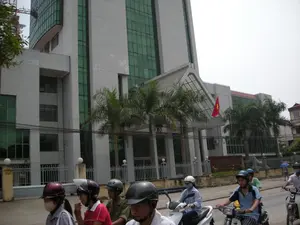 Bảng hiệu màu đỏ trước tòa nhà có ghi: 'VIỆN KINH TẾ VIỆT NAM'. Bối cảnh là một con đường ở thành phố, phía trước một tòa nhà văn phòng lớn, hiện đại. Tòa nhà này có tường trắng và cửa sổ kính màu xanh lá, được xác định là Viện Kinh tế Việt Nam qua bảng hiệu. Một lá cờ tổ quốc Việt Nam được treo gần lối vào. Dọc theo vỉa hè có trồng cây cọ và một hàng rào. Ở tiền cảnh, nhiều người đang tham gia giao thông trên đường bằng xe máy. Tất cả mọi người đều đội mũ bảo hiểm, và một số người đeo khẩu trang. Ở phía xa bên trái, có thể nhìn thấy một cần cẩu xây dựng, cho thấy sự phát triển của khu vực.。（日本語訳）。建物の前にある赤い看板には「VIỆN KINH TẾ VIỆT NAM」と書かれています。背景は、近代的な大きなオフィスビルの前にある、街の通りです。この建物は白い壁と緑色のガラス窓があり、看板によってベトナム経済研究所であると特定されます。入り口の近くにベトナムの国旗が掲げられています。歩道に沿ってヤシの木が植えられ、柵があります。前景では、多くの人々がバイクで道路を通行しています。全員がヘルメットを着用しており、何人かはマスクをしています。遠くの左側には建設用クレーンが見え、この地域の発展を示しています。