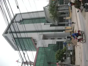 Bức ảnh chụp một con đường ở thành phố, nổi bật là một tòa nhà cao tầng hiện đại với tường trắng và cửa sổ kính màu xanh lá cây. Bầu trời bị che khuất bởi một mạng lưới dây điện chằng chịt. Trên đường phố, có người đang đi xe máy và người đi bộ trên vỉa hè. Một người đàn ông mặc áo xanh dương đang lái một chiếc xe máy màu đỏ. Một người khác mặc áo màu xanh rêu, đội mũ bảo hiểm, cũng đang ngồi trên xe máy. Ở phía xa, có thể thấy một chiếc cần cẩu xây dựng với lá cờ Việt Nam. Có một cột biển báo trên vỉa hè, nhưng chữ viết trên đó không thể đọc rõ, chỉ có thể thấy một tấm biển nhỏ ghi số '475'.。（日本語訳）。都市の通りを写した写真で、白い壁と緑色のガラス窓を持つ近代的な高層ビルが際立っています。空は、入り組んだ電線の網によって覆われています。通りには、バイクに乗っている人や歩道を歩いている人がいます。青いシャツを着た男性が赤いバイクを運転しています。別の人はモスグリーンのシャツを着てヘルメットをかぶり、同じくバイクに乗っています。遠くには、ベトナム国旗を掲げた建設用クレーンが見えます。