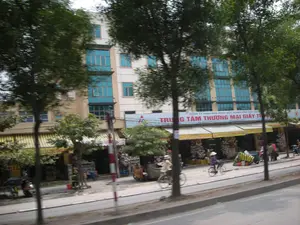 Bối cảnh là một con phố ở Hà Nội, được chụp từ một phương tiện đang di chuyển nên hình ảnh hơi mờ. Dọc theo vỉa hè là hàng cây xanh. Phía sau là một tòa nhà nhiều tầng hiện đại với mặt tiền màu be và nhiều cửa sổ kính màu xanh. Tầng trệt của tòa nhà là một dãy cửa hàng có mái hiên kẻ sọc vàng trắng. Một tấm biển lớn phía trên các cửa hàng có ghi dòng chữ "CÔNG TY TNHH NHÀ NƯỚC MỘT THÀNH VIÊN GIẤY TH..." và bên dưới là dòng chữ lớn hơn "TRUNG TÂM THƯƠNG MẠI GIẤY TH...". Trên đường, có hai người đang đi xe đạp, trong đó có một người đội nón lá. Một số người khác đang đi bộ trên vỉa hè gần các cửa hàng. Có xe máy đậu ven đường. Khung cảnh thể hiện nhịp sống thường nhật trên một con phố thương mại.。（日本語訳）。背景はハノイのある通りで、移動中の乗り物から撮影されたため、画像は少しぼやけている。歩道に沿って緑の並木がある。後ろには、ベージュ色のファサードと多くの青いガラス窓を持つ近代的な高層ビルがある。ビルの1階は、黄と白の縞模様のひさしがある店舗が並んでいる。店舗の上にある大きな看板には、「CÔNG TY TNHH NHÀ NƯỚC MỘT THÀNH VIÊN GIẤY TH...」という文字が書かれており、その下にはより大きな文字で「TRUNG TÂM THƯƠNG MẠI GIẤY TH...」と書かれている。路上には自転車に乗っている人が2人おり、そのうち1人はノンラーをかぶっている。他の何人かの人々が店の近くの歩道を歩いている。道路脇にはバイクが停まっている。この光景は、商業的な通りの日常の営みを示している。
