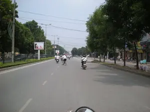 Đây là khung cảnh trên một con đường lớn, có nhiều làn xe tại Hà Nội. Con đường có dải phân cách cứng ở giữa được trồng cây xanh và có hàng rào. Rất nhiều người đang di chuyển trên đường, chủ yếu bằng xe máy, và có một người đi xe đạp. Nhiều người trong số họ mặc áo sơ mi trắng. Hai bên đường và dải phân cách được trồng nhiều cây xanh. Bên phải là dãy các cửa hàng, phía trước có một số xe máy đang đỗ. Có một biển quảng cáo lớn ở dải phân cách bên trái, dường như quảng cáo cho sản phẩm chăm sóc cá nhân của nhãn hiệu 'Dove', nhưng các chi tiết khác không rõ. Các biển hiệu của cửa hàng bên phải cũng không thể đọc được. Bức ảnh được chụp từ góc nhìn của một người đang lái xe máy, có thể thấy một phần gương chiếu hậu ở phía dưới.。（日本語訳）。これはハノイの多車線ある大通りでの光景です。道路には中央に緑の木が植えられ、柵が設置された固定式の中央分離帯があります。多くの人々が道路上を移動しており、主にバイクを利用していますが、自転車に乗っている人も一人います。彼らの多くは白いシャツを着ています。道路の両側と中央分離帯には多くの緑の木が植えられています。右側には店舗が並んでおり、その前には数台のバイクが駐車しています。左側の中央分離帯には大きな広告看板があり、「Dove」ブランドのパーソナルケア製品を宣伝しているようですが、その他の詳細は不明です。右側の店舗の看板も読み取れません。この写真はバイクを運転している人の視点から撮影されたもので、下部にバックミラーの一部が見えます。