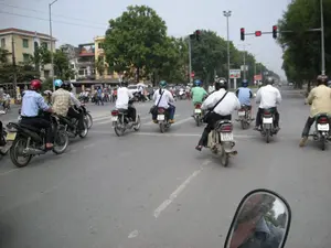 Bối cảnh là một con đường nhựa rộng lớn trong một thành phố, có nhiều làn xe. Rất đông người đang tham gia giao thông, chủ yếu bằng xe máy. Họ đang dừng lại tại một ngã tư để chờ đèn đỏ. Đèn tín hiệu giao thông phía trước đang hiển thị màu đỏ và đồng hồ đếm ngược chỉ số '00'. Nhiều người điều khiển xe máy đội mũ bảo hiểm với các màu sắc khác nhau như đỏ và xanh dương. Trang phục của họ là quần áo thường ngày như áo sơ mi và quần dài. Hai bên đường là hàng cây xanh lớn và các tòa nhà cao tầng. Ở phía xa có một vài tấm biển quảng cáo nhưng không thể đọc rõ nội dung. Phía dưới cùng của bức ảnh là một phần gương chiếu hậu của chiếc xe mà người chụp ảnh đang ngồi trên. Khung cảnh cho thấy một hoạt động giao thông đông đúc và quen thuộc vào ban ngày trên một con phố ở Hà Nội.。（日本語訳）。背景は、ある都市の、多くの車線がある広いアスファルトの道路です。非常に多くの人々が、主にバイクで交通に参加しています。彼らは赤信号を待つために交差点で停止しています。前方の交通信号機は赤色を表示しており、カウントダウンタイマーは「00」を示しています。多くのバイク運転手は、赤や青など様々な色のヘルメットをかぶっています。彼らの服装は、シャツや長ズボンのような普段着です。道路の両側には、大きな緑の並木と高層ビルがあります。遠方にはいくつかの広告看板がありますが、内容ははっきりと読み取れません。この光景は、ハノイのある通りでの日中の、混雑した見慣れた交通の様子を示しています。