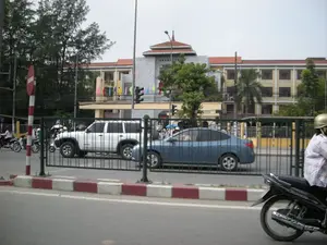 Đây là một cảnh đường phố tại Hà Nội. Ở phía sau, có một tòa nhà lớn, nhiều tầng với kiến trúc kết hợp giữa hiện đại và truyền thống, có thể là một cơ quan công quyền hoặc một tòa nhà công cộng. Tòa nhà được sơn màu vàng và xám, trên ban công có treo nhiều lá cờ nhỏ nhiều màu sắc. Dọc theo con đường là dòng xe cộ đang lưu thông, bao gồm một chiếc ô tô SUV màu trắng và một chiếc sedan màu xanh dương phía sau hàng rào sắt. Nhiều người đang điều khiển xe máy, một phương tiện giao thông phổ biến. Ở góc dưới bên phải, một người đang lái xe máy và đội mũ bảo hiểm. Bên lề đường có một cột biển báo giao thông sọc đỏ trắng, trên đó có biển báo hình tròn nền đỏ, vạch ngang màu trắng ở giữa, là biển báo cấm đi ngược chiều. Ngoài ra còn có đèn tín hiệu giao thông, cây xanh và vỉa hè được sơn sọc đỏ trắng.。（日本語訳）。これはハノイのある街の風景です。後方には、現代建築と伝統建築を組み合わせた大きな多層階の建物があり、官公庁か公共の建物かもしれません。建物は黄色と灰色で塗られており、バルコニーには色とりどりの小さな旗がたくさん飾られています。道路沿いでは車が行き交っており、白いSUVと鉄柵の後ろに青いセダンが見えます。多くの人が、一般的な交通手段であるバイクを運転しています。右下隅では、ヘルメットをかぶった人がバイクを運転しています。道端には赤と白の縞模様の交通標識のポールがあり、その上には赤地に中央に白い横線が入った円形の標識、つまり進入禁止の標識が設置されています。その他にも、信号機、緑の木々、赤と白の縞模様に塗られた歩道があります。