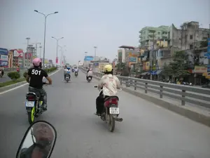 Bức ảnh chụp một cảnh giao thông trên một con đường trên cao hoặc cầu vượt ở Hà Nội. Nhiều người đang đi xe máy trên đường, tất cả đều đội mũ bảo hiểm. Ở phía trước, một người đàn ông mặc áo phông đen in số "501" và đội mũ bảo hiểm màu hồng. Bên cạnh anh ta, một người khác mặc áo hoa và đội mũ bảo hiểm màu vàng đang lái một chiếc xe máy màu đỏ. Phía xa hơn có nhiều xe máy khác đang lưu thông. Bên trái đường là dải phân cách có trồng cây xanh. Bên phải là lan can bê tông, phía sau là các dãy nhà phố nhiều tầng san sát nhau với nhiều biển hiệu và biển quảng cáo. Một số chữ trên biển quảng cáo có thể đọc được như "LAPTOP" và "Midea". Phía dưới bên trái bức ảnh là hình ảnh phản chiếu của người chụp trong gương chiếu hậu của xe máy.。（日本語訳）。写真はハノイの高架道路または陸橋での交通風景を撮影したものです。多くの人々が道路でバイクに乗っており、全員がヘルメットを着用しています。前方には、「501」という数字がプリントされた黒いTシャツを着て、ピンク色のヘルメットをかぶった男性がいます。彼の隣では、花柄のシャツを着て黄色のヘルメットをかぶった別の人が、赤いバイクを運転しています。さらに遠くには、他にも多くのバイクが走行しています。道路の左側には、緑の木が植えられた中央分離帯があります。右側にはコンクリートの欄干があり、その後ろには多くの看板や広告がある多層階の町家が密集しています。広告看板のいくつかの文字は、「LAPTOP」や「Midea」のように読み取ることができます。写真の左下には、バイクのバックミラーに映った撮影者の姿があります。