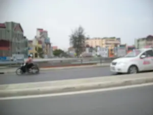 Bức ảnh bị mờ, ghi lại một cảnh giao thông trên một con đường hoặc cây cầu ở khu vực đô thị, có thể là ở Hà Nội. Con đường có dải phân cách và lan can. Ở phía bên trái, một người đội mũ bảo hiểm đang lái một chiếc xe máy. Ở phía bên phải, một chiếc ô tô màu trắng, có khả năng là xe taxi, đang di chuyển. Trên thân xe taxi có các họa tiết và chữ màu hồng nhưng không thể đọc rõ do ảnh mờ. Phía sau là khung cảnh các tòa nhà nhiều tầng san sát nhau. Bầu trời có màu xám trắng.。（日本語訳）。ぼやけた写真で、都市部、おそらくハノイにある道路か橋の上の交通風景を記録したものである。道路には中央分離帯とガードレールがある。左側では、ヘルメットをかぶった人がバイクを運転している。右側では、白い自動車、おそらくタクシーと思われる車が走行している。タクシーの車体にはピンク色の模様と文字があるが、写真がぼやけているためはっきりと読み取ることはできない。後方には、高層ビルが密集している光景が広がっている。空は灰白色である。