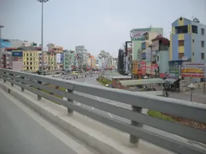 Đây là một khung cảnh đường phố ở Hà Nội, được chụp từ trên một cây cầu vượt. Con đường nhựa rộng và uốn cong, có dải phân cách trồng cỏ ở giữa. Trên đường có rất nhiều người đang đi xe máy và một chiếc xe buýt màu vàng. Hai bên đường là những dãy nhà ống cao tầng, nhiều màu sắc, san sát nhau, đặc trưng của kiến trúc đô thị Việt Nam. Các tòa nhà được bao phủ bởi vô số biển hiệu quảng cáo với nhiều kích cỡ và màu sắc khác nhau. Mặc dù hầu hết các chữ đều khó đọc do khoảng cách, có thể nhận ra một tấm biển lớn màu đỏ và vàng ghi 'BIA HƠI HIẾU BÉO'. Khung cảnh tổng thể cho thấy giao thông và sinh hoạt thường ngày trên một con phố sầm uất của thành phố.。（日本語訳）。これはハノイのある通りの風景で、陸橋の上から撮影されたものです。アスファルトの道は広くカーブしており、中央には草が植えられた分離帯があります。道路にはバイクに乗っている多くの人々と、一台の黄色いバスがいます。道路の両側には、ベトナムの都市建築に特徴的な、色とりどりの高層のチューブハウスが密集して並んでいます。建物は、さまざまなサイズと色の無数の広告看板で覆われています。ほとんどの文字は距離があるため読みづらいですが、「BIA HƠI HIẾU BÉO」と書かれた赤と黄色の大きな看板が認識できます。全体的な風景は、都市の賑やかな通りでの日常的な交通と生活を示しています。