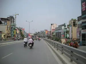 Bức ảnh ghi lại cảnh giao thông trên một con đường trên cao hoặc cầu vượt tại một khu vực đô thị ở Hà Nội. Nhiều người đang điều khiển xe máy, phương tiện di chuyển phổ biến, trên một con đường rộng rãi có dải phân cách và lan can. Hai bên đường là những dãy nhà ống cao tầng, san sát nhau với nhiều màu sắc. Phía xa bên trái, một công trình xây dựng đang dang dở với một chiếc cần cẩu lớn. Có rất nhiều biển hiệu quảng cáo. Một số biển hiệu có thể đọc được chữ như: "TÂY SƠN", "CAFE", "HOTEL", "Nhật Linh" ở phía bên trái, và một biển hiệu dọc lớn ghi "KHUÔNG VIÊN" ở phía bên phải. Một lá cờ Việt Nam cũng được treo trên một tòa nhà. Khung cảnh cho thấy sự phát triển và nhịp sống sôi động của một thành phố Việt Nam.。（日本語訳）。写真は、ハノイのある都市部における高架道路または陸橋の上の交通風景を捉えている。多くの人々が、一般的な交通手段であるバイクを、中央分離帯とガードレールのある広々とした道路で運転している。道路の両側には、様々な色の高層チューブハウスが密集して建ち並んでいる。広告看板がたくさんある。いくつかの看板は文字が読め、左側には「TÂY SƠN」、「CAFE」、「HOTEL」、「Nhật Linh」などがあり、右側には「KHUÔNG VIÊN」と書かれた大きな縦長の看板がある。この風景は、あるベトナムの都市の発展と活気ある生活リズムを示している。