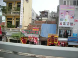Bức ảnh bị mờ, chụp lại một khung cảnh đường phố ở một khu đô thị tại Việt Nam, có khả năng là Hà Nội. Các tòa nhà cao tầng, kiểu nhà ống, được xây san sát nhau. Bên trái là một tòa nhà sơn màu vàng với ban công, trên đó có treo một biển hiệu dọc màu trắng với các chữ cái không rõ ràng, có thể là 'NGOC VIEN'. Ở trung tâm, có một cửa hàng với biển hiệu màu xanh lá và đỏ mang logo 'FUJIFILM', cho thấy đây là một tiệm ảnh. Phía trên cửa hàng này là một mái nhà lợp ngói đỏ, một phần được che bằng tấm bạt màu xanh dương. Bên phải là một tòa nhà màu xám với một tấm biển quảng cáo lớn màu hồng có hình một người phụ nữ, bên cạnh có chữ 'In' (in ấn). Phía trước các cửa hàng được trang trí bằng các dải băng, biểu ngữ màu đỏ và vàng, cùng những vật trang trí trông giống đèn lồng, gợi ý không khí lễ hội hoặc chuẩn bị cho một dịp đặc biệt. Phần tiền cảnh có một lan can màu xám, cho thấy ảnh được chụp từ một vị trí trên cao như cầu hoặc cầu vượt.。（日本語訳）。写真はぼやけており、ベトナムのある都市部、おそらくハノイの街の風景を撮影したものである。チューブハウス様式の高層ビルが互いに密集して建てられている。中央には、「FUJIFILM」のロゴが入った緑と赤の看板を掲げた店があり、これが写真店であることを示している。この店の上には赤い瓦屋根があり、その一部は青い防水シートで覆われている。右側には灰色の建物があり、そこには女性の絵が描かれた大きなピンク色の広告看板があり、その横には「In」（印刷）という文字がある。店の前は、赤と黄色のリボンや横断幕、そして提灯のような装飾品で飾られており、祭りの雰囲気や特別な行事の準備を示唆している。前景には灰色の手すりがあり、写真が橋や陸橋のような高い場所から撮影されたことを示している。