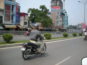 Bức ảnh chụp một con đường phố đông đúc ở Hà Nội vào ban ngày. Con đường rộng, có nhiều làn xe và có dải phân cách trồng các bụi cây nhỏ. Hoạt động chính là giao thông, với nhiều người đang điều khiển xe máy trên đường. Người đàn ông ở phía trước đang lái xe máy và đội mũ bảo hiểm.。Hai bên đường là những dãy nhà ống cao tầng san sát nhau, là kiến trúc đặc trưng của đô thị Việt Nam, với các cửa hàng kinh doanh ở tầng trệt. Có rất nhiều biển hiệu quảng cáo, một số có thể đọc được:。- Biển hiệu lớn màu đỏ và xanh dương trên một tòa nhà: "Thẩm mỹ viện Nhật Linh".。- Biển hiệu màu xanh dương trên tòa nhà màu xanh lam: "PHÚC ÂN", bên dưới có ghi địa chỉ "ĐC: 182 TÂY SƠN - ĐỐNG ĐA - HÀ NỘI".。- Biển hiệu màu vàng có chữ "TÂY SƠN".。- Biển hiệu ở phía xa bên trái: "NỘI THẤT GIA ĐÌNH" và "ĐÓNG GIƯỜNG TỦ BÀN GHẾ".。- Các biển hiệu khác có nội dung như "CÔNG TY TNHH..." và "CÔNG TY CP ĐẦU TƯ VÀ...".。Ở phía sau, có một tòa nhà cao tầng đang trong quá trình xây dựng và một chiếc cần cẩu.。（日本語訳）。日中のハノイの混雑した街路を写した写真です。道は広く、多くの車線があり、小さな低木が植えられた中央分離帯があります。主な活動は交通で、多くの人々が路上でバイクを運転しています。前方にいる男性はバイクを運転し、ヘルメットをかぶっています。道の両側には、ベトナムの都市に特徴的な建築様式である、高層のチューブハウスがびっしりと並んでおり、1階には商業店舗があります。広告看板がたくさんあり、そのうちいくつかは読み取れます：。- ある建物にある赤と青の大きな看板：「Thẩm mỹ viện Nhật Linh」（ニャットリン美容院）。- 青い建物にある青い看板：「PHÚC ÂN」（フックアン）、その下には「ĐC: 182 TÂY SƠN - ĐỐNG ĐA - HÀ NỘI」（住所：ハノイ市ドンダー区タイソン通り182番地）と住所が記載されています。- 「TÂY SƠN」（タイソン）と書かれた黄色い看板。- 左奥の看板：「NỘI THẤT GIA ĐÌNH」（家庭用家具）と「ĐÓNG GIƯỜNG TỦ BÀN GHẾ」（ベッド・タンス・テーブル・椅子の製作）。- その他の看板には「CÔNG TY TNHH...」（有限会社...）や「CÔNG TY CP ĐẦU TƯ VÀ...」（投資...株式会社）といった内容があります。後方には、建設中の高層ビルと一台のクレーンがあります。