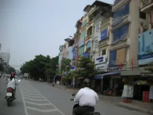 Bức ảnh chụp một cảnh đường phố ở một khu đô thị, có khả năng là Hà Nội. Bên phải đường là một dãy nhà ống cao tầng san sát nhau, một kiểu kiến trúc phổ biến ở Việt Nam. Các tòa nhà này được sử dụng làm cả nhà ở và cửa hàng kinh doanh ở tầng trệt. Có rất nhiều biển hiệu và biển quảng cáo được treo trên mặt tiền các tòa nhà, nhưng chữ viết trên đó bị mờ và không thể đọc được. Trên đường, nhiều người đang tham gia giao thông bằng xe máy. Ở phía trước, có một người đàn ông mặc áo trắng và một người phụ nữ mặc áo trắng, đội mũ bảo hiểm màu đỏ đang điều khiển xe máy. Dọc theo con đường có cây xanh. Con đường được trải nhựa và có vạch kẻ đường. Toàn bộ khung cảnh cho thấy một ngày sinh hoạt bình thường trên đường phố Việt Nam.。（日本語訳）。写真は、ある都市部の街の風景を写したもので、ハノイである可能性があります。道路の右側には、ベトナムで一般的な建築様式である、高層のチューブハウスが密集して並んでいます。これらの建物は、住居と1階の店舗の両方として使用されています。建物の正面には多くの看板や広告が掲げられていますが、そこに書かれた文字はぼやけていて読むことができません。道路では、多くの人々がバイクで交通に参加しています。前方には、白いシャツを着た男性と、白いシャツを着て赤いヘルメットをかぶった女性がバイクを運転しています。道沿いには緑の木々があります。道路は舗装されており、車線が引かれています。全体の光景は、ベトナムの路上での普通の日常生活の一日を示しています。