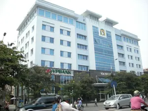 Bức ảnh chụp một tòa nhà văn phòng hiện đại, cao tầng trên một con phố đô thị. Tòa nhà có mặt tiền sơn màu trắng và xanh nhạt với nhiều cửa sổ kính. Trên tầng trệt của tòa nhà có một biển hiệu lớn màu đỏ và xanh lá cây với dòng chữ 'VPBANK' và bên dưới là 'NGÂN HÀNG NGOÀI QUỐC DOANH'. Bên cạnh đó, phía trên một lối vào khác có biển hiệu ghi 'TÒA NHÀ' nhưng phần chữ còn lại không thể đọc rõ. Phía trên cao của tòa nhà có một logo màu xanh lá cây và vàng. Khung cảnh đường phố cho thấy hoạt động giao thông với nhiều xe máy và ô tô đang di chuyển. Một số người đang đi xe máy và đội mũ bảo hiểm. Cây xanh được trồng dọc theo vỉa hè.。（日本語訳）。写真は、都市の通りにある近代的な高層オフィスビルを写しています。ビルのファサードは白と水色に塗られており、ガラス窓がたくさんあります。ビルの1階には、赤と緑の大きな看板があり、「VPBANK」という文字と、その下に「NGÂN HÀNG NGOÀI QUỐC DOANH」と書かれています。その隣、別の入り口の上には「TÒA NHÀ」と書かれた看板がありますが、残りの文字ははっきりと読み取れません。ビルの上部には、緑と黄色のロゴがあります。街の風景は、多くのバイクや車が移動している交通の様子を示しています。何人かの人々がバイクに乗り、ヘルメットをかぶっています。歩道に沿って緑の木が植えられています。