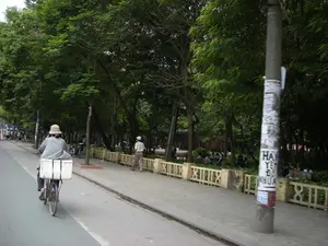 Trên cột điện ở bên phải có dán rất nhiều tờ rơi, quảng cáo. Một số chữ có thể đọc được một phần ở phía dưới là 'HA', 'YET', 'ĐỂ', 'NHỰA', nhưng toàn bộ nội dung không thể nhận dạng rõ.。Bối cảnh là một con đường phố rợp bóng cây, có thể là ở Hà Nội. Một người đội mũ rộng vành đang đạp một chiếc xe đạp trên đường, phía sau xe có chở một chiếc thùng lớn màu trắng, có thể là để vận chuyển hàng hóa. Trên vỉa hè, một người đàn ông đang đi bộ. Bên trong hàng rào bê tông thấp là một khu vực giống công viên với nhiều cây xanh, nơi có một vài nhóm người đang ngồi nghỉ ngơi, trò chuyện. Phía xa trên đường có thể thấy thêm người và xe máy đang di chuyển.。（日本語訳）。右側の電柱には、たくさんのチラシや広告が貼られています。下の方で部分的に読み取れる文字は「HA」、「YET」、「ĐỂ」、「NHỰA」ですが、全体の内容ははっきりと識別できません。背景は木陰の多い街路で、おそらくハノイでしょう。つばの広い帽子をかぶった人が道路で自転車を漕いでおり、自転車の後ろには大きな白い箱を積んでいます。これは商品を運ぶためのものかもしれません。歩道では、一人の男性が歩いています。低いコンクリートの柵の内側は、たくさんの緑の木々がある公園のようなエリアで、いくつかのグループの人々が座って休憩したり、おしゃべりしたりしています。道路の遠くには、さらに人々やバイクが移動しているのが見えます。