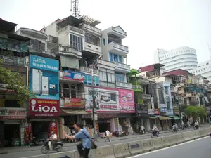 Bức ảnh chụp một con phố sầm uất ở Hà Nội với những dãy nhà ống cao tầng, san sát nhau, đây là kiến trúc đặc trưng của đô thị Việt Nam. Tầng trệt của các ngôi nhà là các cửa hàng kinh doanh. Trên đường có nhiều người đang di chuyển bằng xe máy, và có một dải phân cách bằng bê tông ở giữa đường. Có rất nhiều biển quảng cáo với các nội dung có thể đọc được như sau: Một biển quảng cáo lớn màu xanh của thương hiệu 'LiOA' với khẩu hiệu 'Giữ gìn tuổi thọ Thiết bị điện'. Ngay bên dưới là biển hiệu màu đỏ của 'CÔNG TY ỔN ÁP NHẬT LINH', thương hiệu 'LIOA' và dòng chữ 'Điểm và giới thiệu sản phẩm'. Một cửa hàng bên trái có biển ghi địa chỉ '279 TÂY SƠN ĐT: 8574153'. Các biển hiệu khác có thể nhìn thấy bao gồm 'ĐỆM CAO CẤP', 'ĐẠI LÝ BÌNH NÓNG LẠNH', và 'EVERON'. Trên nóc một tòa nhà có một cột tháp viễn thông. Ở phía xa là một tòa nhà văn phòng hiện đại màu trắng.。（日本語訳）。この写真はハノイの賑やかな通りを撮影したもので、高層のチューブハウスが密集して立ち並んでおり、これはベトナムの都市に特有の建築様式です。家々の1階は店舗になっています。道路上では多くの人々がバイクで移動しており、道路の中央にはコンクリート製の中央分離帯があります。以下のように読み取れる内容の広告看板がたくさんあります。「LiOA」ブランドの大きな青い広告看板には、「電気製品の寿命を守る」というスローガンが書かれています。そのすぐ下には、「CÔNG TY ỔN ÁP NHẬT LINH」（ニャットリン変圧器会社）の赤い看板があり、「LIOA」ブランドと「製品紹介・販売拠点」という文字が書かれています。左側のある店には、「279 TÂY SƠN ĐT: 8574153」という住所が書かれた看板があります。ある建物の屋上には通信塔が立っています。遠くには近代的な白いオフィスビルが見えます。