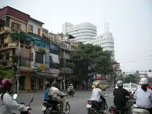 Cảnh quan một con đường phố đông đúc ở Hà Nội. Dòng phương tiện giao thông chủ yếu là xe máy, với nhiều người đang điều khiển xe trên đường, tất cả đều đội mũ bảo hiểm. Một số xe chở hai người. Phía xa có một chiếc xe buýt nhỏ màu vàng và một chiếc taxi màu trắng. Bên trái đường là một dãy nhà ống nhiều tầng kiểu cũ, san sát nhau, tầng trệt được sử dụng làm cửa hàng. Có rất nhiều biển hiệu quảng cáo treo trên các tòa nhà, trong đó có một biển hiệu ghi chữ 'AOC', các biển hiệu khác chữ viết không thể đọc rõ. Phía sau những ngôi nhà cũ là một cây xanh lớn và một tòa nhà văn phòng hiện đại, màu trắng với kiến trúc uốn lượn nổi bật. Bầu trời u ám, và có nhiều dây điện chằng chịt trên đường phố.。（日本語訳）。ハノイの混雑した通りの風景。交通手段は主にバイクで、多くの人が道路で運転しており、全員がヘルメットを着用している。いくつかのバイクは二人乗りである。遠くには黄色い小型バスと白いタクシーが一台ある。道路の左側には、古い様式の多層階建てのチューブハウスが密集して並んでおり、1階は店舗として使用されている。建物には多くの広告看板が掲げられており、その中には「AOC」と書かれた看板が一つあるが、他の看板の文字ははっきりと読み取れない。古い家々の後ろには、大きな緑の木と、際立った曲線的な建築様式の白い近代的なオフィスビルがある。空は曇っており、通りには多くの電線が張り巡らされている。